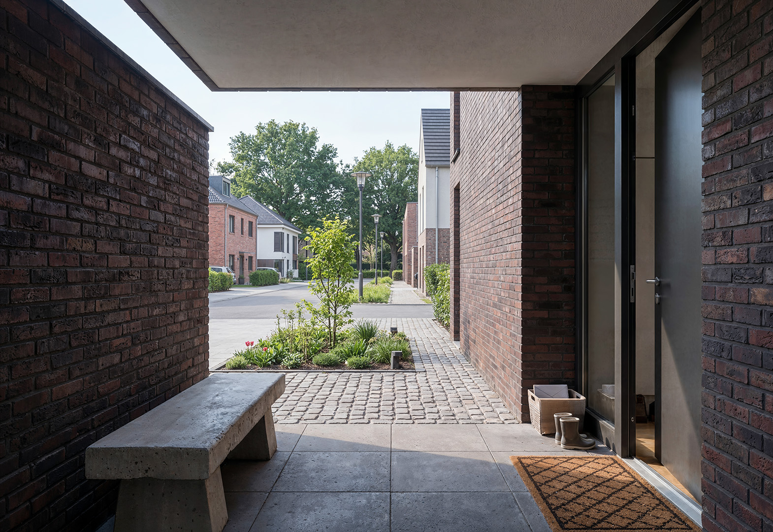 KI-gestützte Architekturvisualisierung: überdachter Eingangsbereich eines Reihenhauses mit dunkelroter Klinkerfassade, Betonbank, anthrazitfarbener Eingangstür und Blick auf begrünten Gemeinschaftsweg mit Kopfsteinpflaster, Neubauquartier Kevelaer