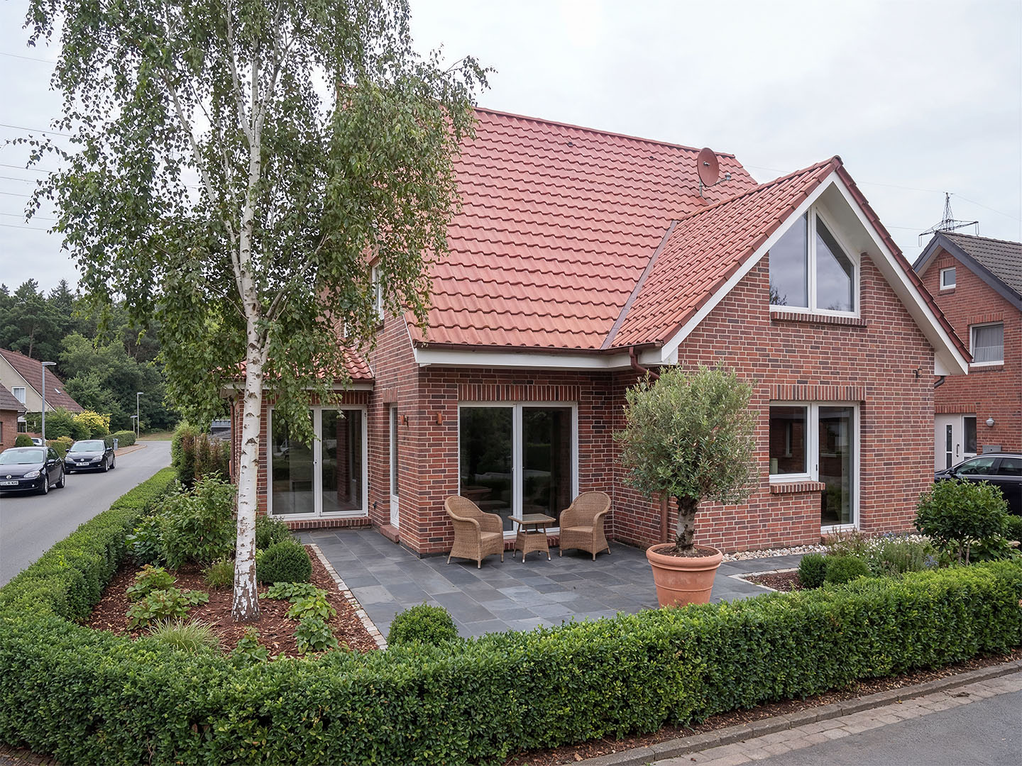 Mittels KI erstelltes Architekturbild: Einfamilienhaus mit roter Klinkerfassade und Satteldach, anthrazitfarbene Terrassenplatten mit Rattanstühlen und Beistelltisch, Olivenbaum im Terrakottatopf, gepflegte Buchshecken und Birke im Vorgarten, Wohnstraße im Hintergrund