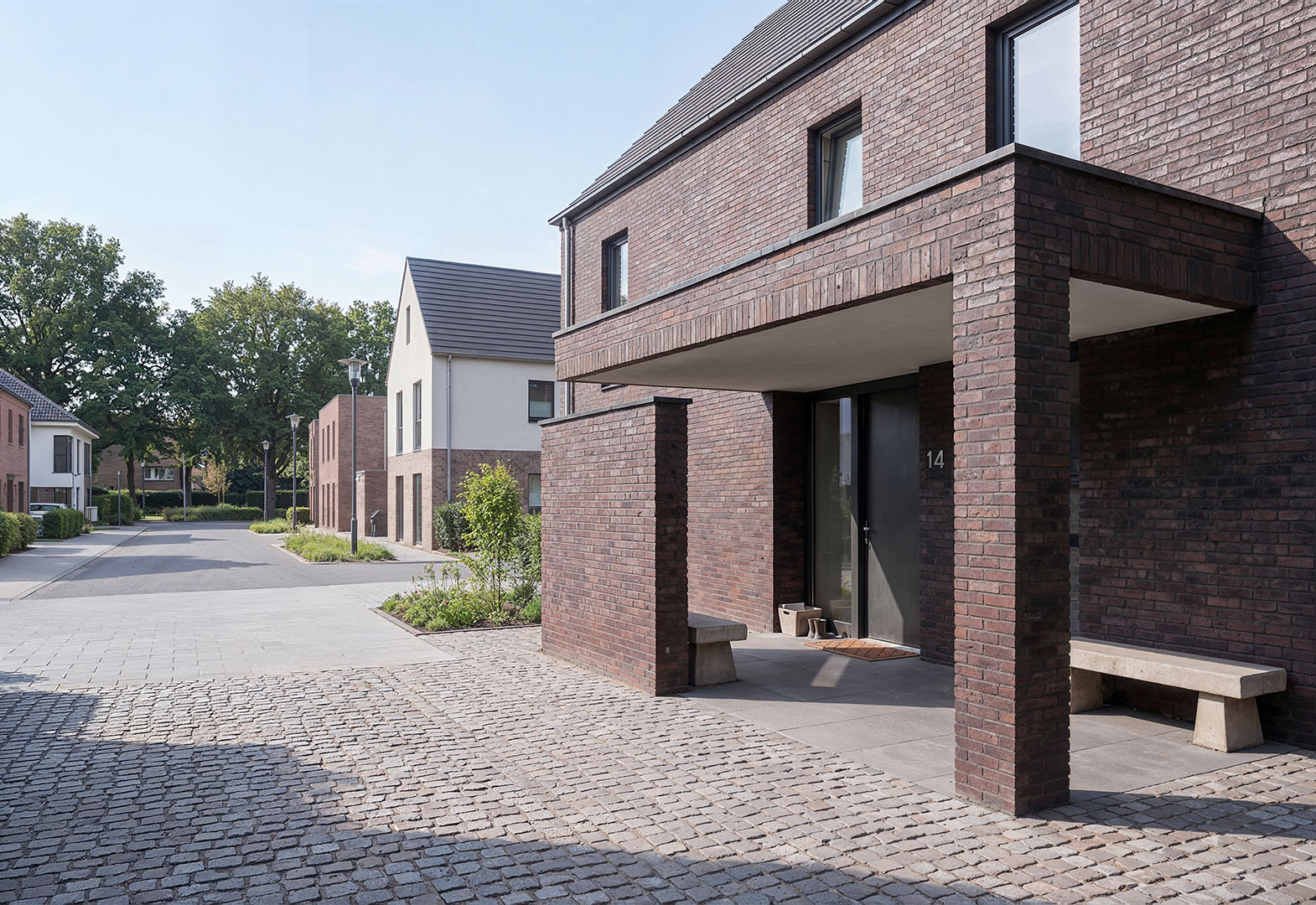 Fotorealistisches Architekturbild: moderne Reihenhaussiedlung mit dunkelroten Klinkerfassaden, auskragendem Vordach über dem Eingang, Betonbank und Kopfsteinpflaster-Gemeinschaftsweg, weitere Wohnhäuser unterschiedlicher Typologie im Hintergrund, Kevelaer am Niederrhein