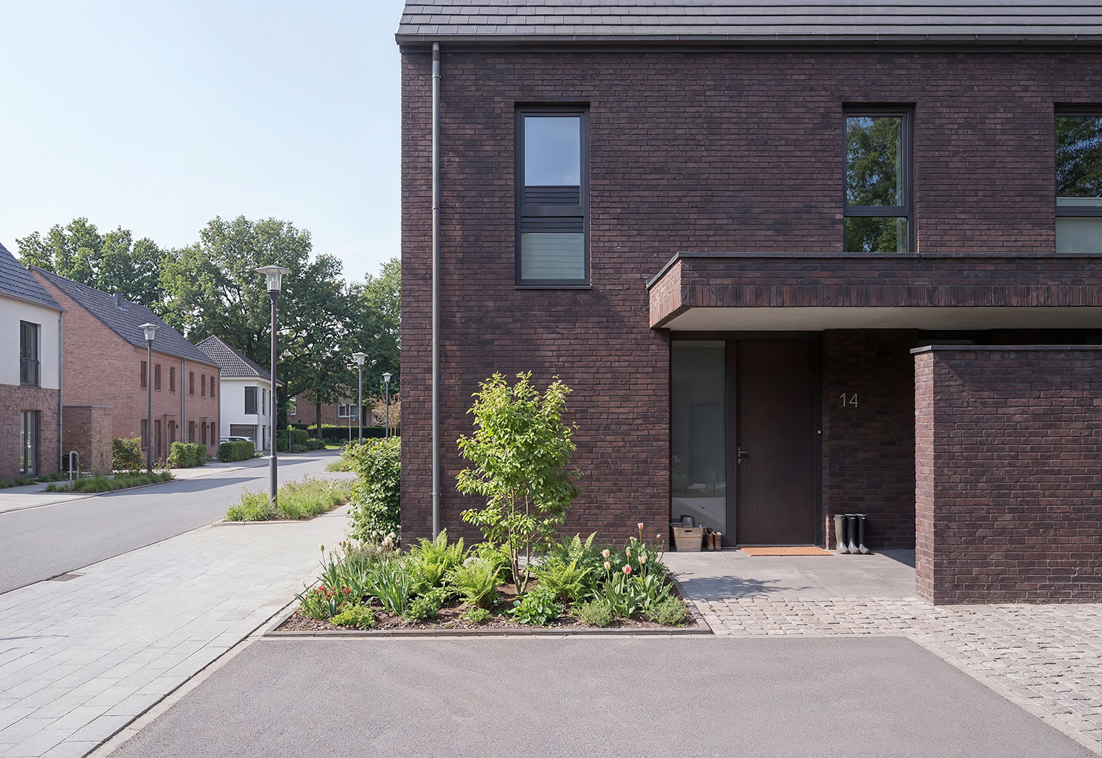 Mittels KI erstelltes Architekturbild: zweigeschossiges Reihenhaus mit dunkelbrauner Klinkerfassade, Balkon, überdachtem Eingangsbereich mit Hausnummer 14, begrüntem Vorgartenbeet mit Tulpen und Farnen, ruhige Wohnstraße im Neubauquartier Kevelaer